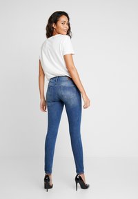 Mager blauwe jeans met een vervaagde uitstraling, gecombineerd met een eenvoudig wit T-shirt. Het model draagt zwarte hoge hakken en kijkt om.