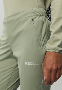 Pantaloni da esterno di un verde oliva chiaro con vita elasticizzata, dettagli con tasche con zip e un logo sulla coscia sinistra. Tessuto di texture morbida.