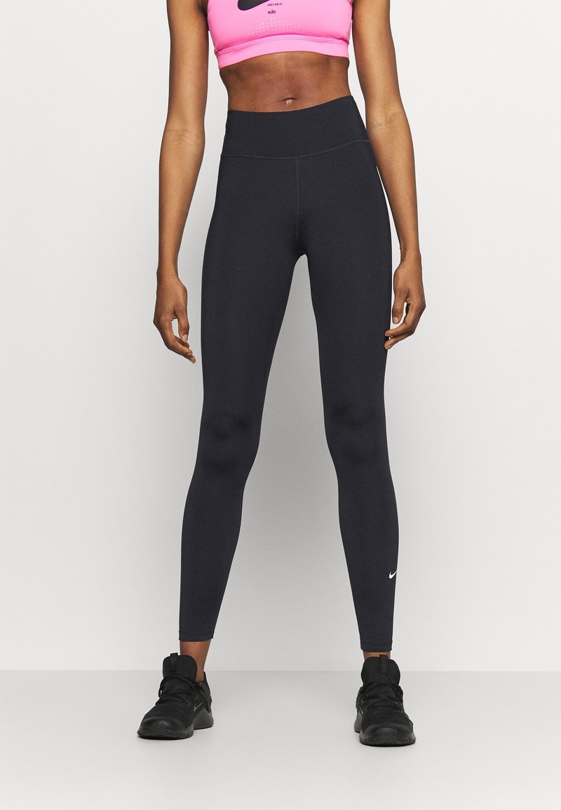Femme portant des leggings noirs taille haute, des baskets noires et un soutien-gorge de sport Nike rose, se tenant contre un fond blanc uni.