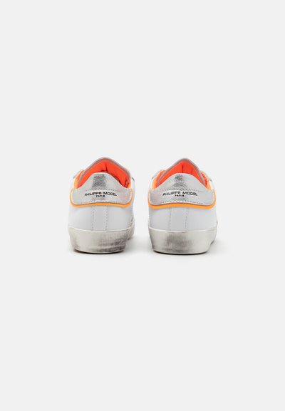 Białe skórzane sneakersy z srebrnym obcasem, pomarańczowymi akcentami i gumową podeszwą. Widok z tyłu pokazuje logo i lekkie zużycie krawędzi podeszwy.