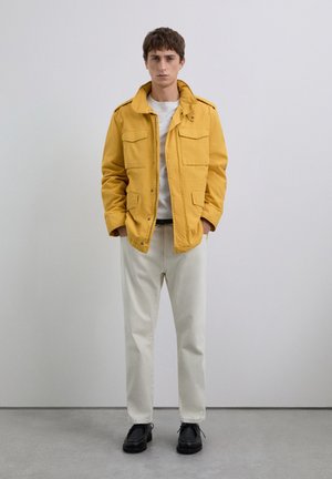 Man in mosterdgele jas over witte shirt, lichtbeige broek en zwarte schoenen staat tegen een effen grijze muur.