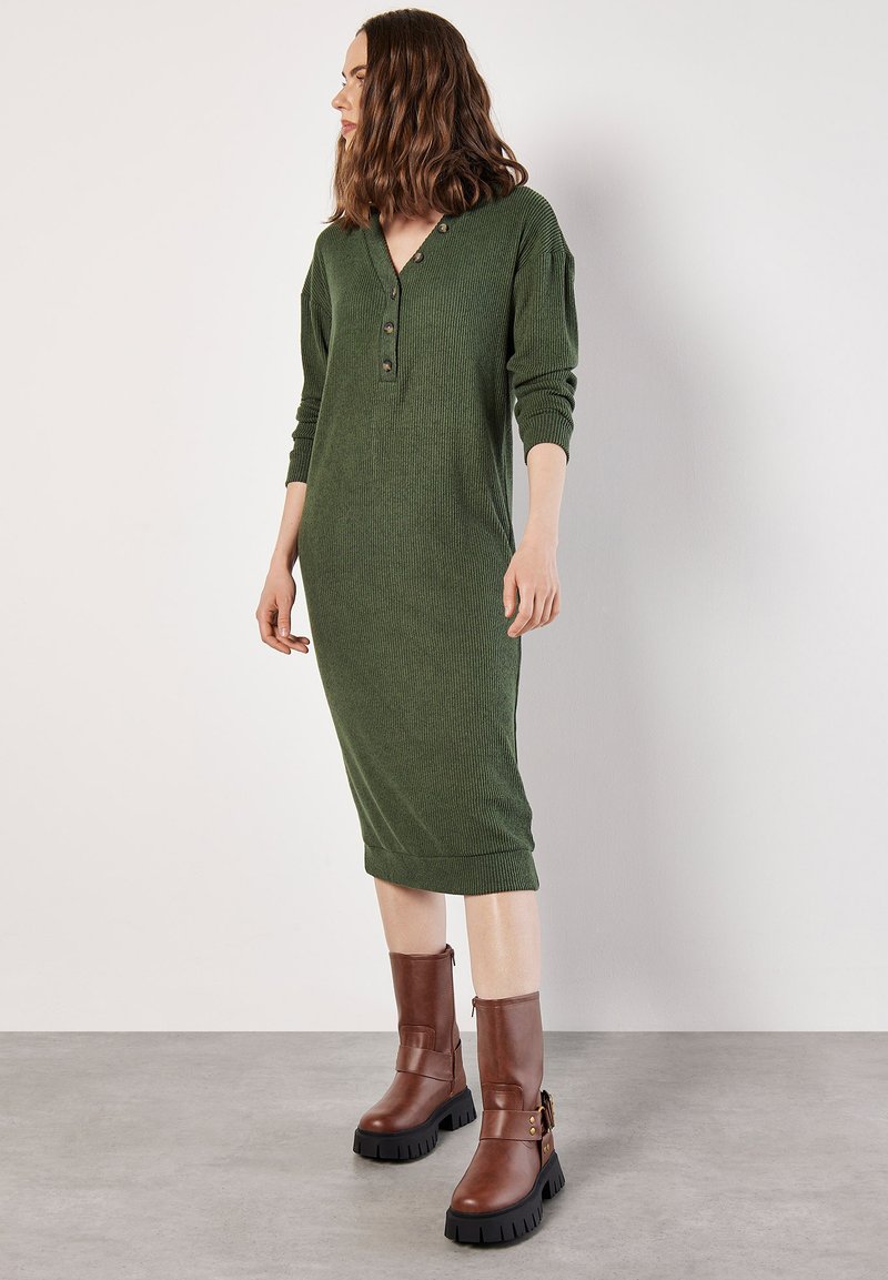 Rippstrickkleid in Grün mit tiefem V-Ausschnitt und Knopfdetails, ergänzt durch braune Ankle Boots mit chunky Sohlen und Akzentriemen.
