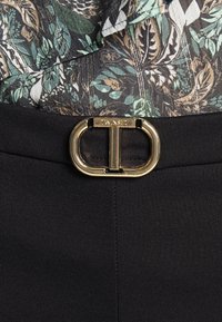 Svarta byxor med ett bälte med guldspränne och en framträdande "T"-design, kombinerat med en mönstrad tygtop i gröna och bruna nyanser.