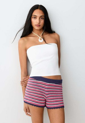 Femme aux longs cheveux noirs portant un top blanc bustier, un short en crochet rayé rose, blanc et marine, et un collier avec un pendentif circulaire.