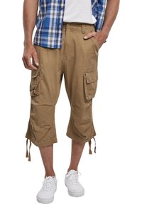 Kaki cargo shorts s više bočnih džepova, podesivim vezicama na rubu i opuštenim krojem, u kombinaciji s bijelim tenisicama.