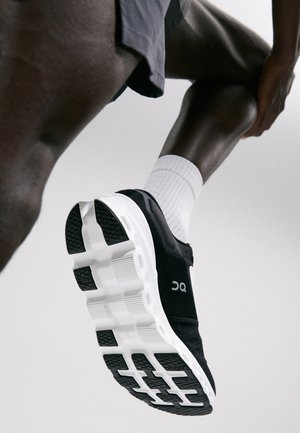 Zapatilla deportiva negra con suela blanca, que presenta un upper texturizado y un logo. La zapatilla tiene acolchado visible y un diseño elegante.