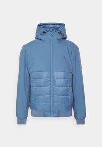 Světle modrá zateplená bunda s kapucí, s předním zipem, elastickými manžetami a strukturovaným panelem přes hrudník. Logo na levé horní straně.