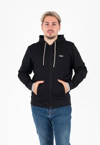 Schwarze Zip-Hoodie aus weichem Stoff, mit beigen Kordeln, Fronttaschen und kontrastierenden weißen Streifen an den Bündchen.