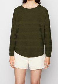 Olivgrüner Strickpullover mit strukturierten Streifen, rundem Ausschnitt und langen Raglanärmeln. Kombiniert mit hellbeigen Shorts.