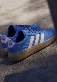 Blue Adidas sneakers with white stripes and gum soles resting on a concrete surface, lit by soft natural light.