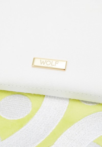 Balts teksturētais maks ar zelta logotipa plāksni, uz kuras ir rakstīts "WOLF ESTD 1834", novietots uz zaļgani pelēka rakstaina fona.