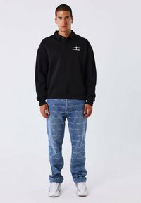 Zwarte langemouwen polo met een logo op de borst, gecombineerd met blauwe gedessineerde denim jeans en witte sneakers.