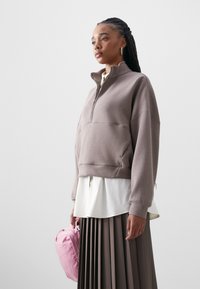 Sweatshirt marrom com fechoZIP e ajuste cropped, usado sobre uma camisa branca em camadas. O modelo segura uma pequena bolsa rosa. Saia cinza plissada visível.