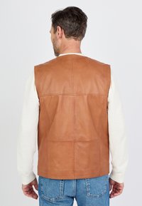 Tan leren vest met een gladde textuur, voorzien van een eenvoudig ontwerp, mouwloos model en twee achterzakken. Draagt over een lichte sweater.