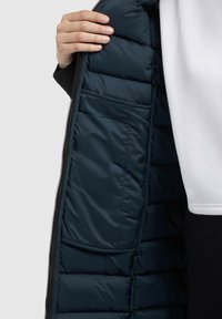 Interieur van een marineblauwe gewatteerde jas, met horizontale quilted inzet, een zak en een zwarte rits. De buitenstof lijkt glad en duurzaam.
