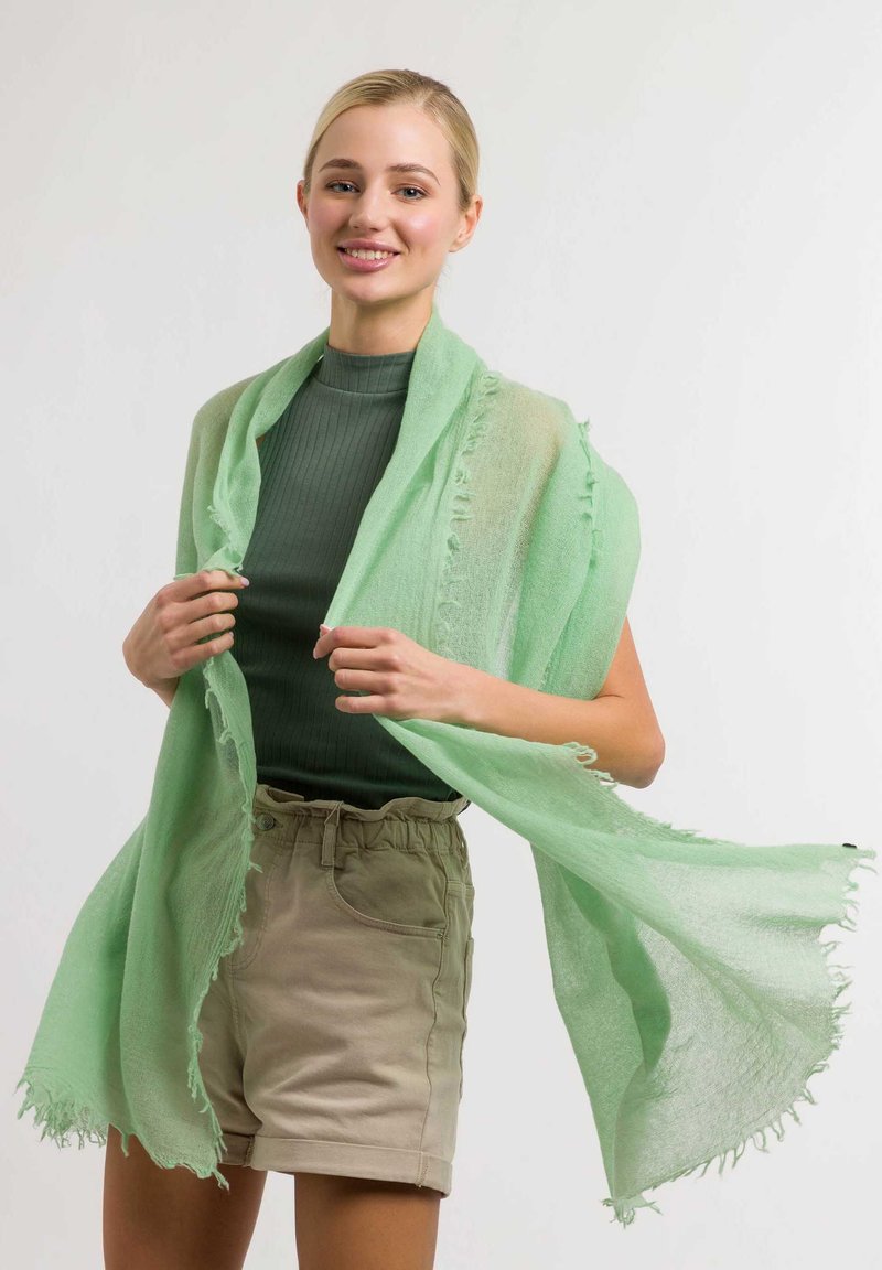Eșarfă verde deschis, cu margini ciufulite, dintr-un material transparent, purtată peste un top verde cu dungi și pantaloni scurți bej. Textură moale și design fluid.