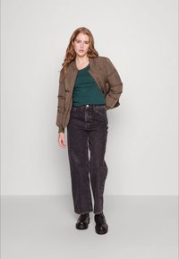 Jaqueta bomber acolchoada castanha, t-shirt azul-turquesa por baixo, jeans pretos de cintura alta e botas pretas chunky. Modelo está de pé sobre um fundo neutro.