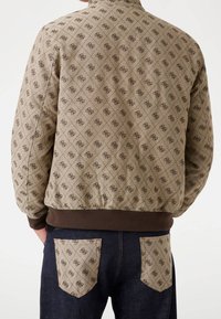 Giacca trapuntata beige con motivo a logo marrone scuro, abbinata a jeans scuri con tasche posteriori beige che presentano lo stesso motivo a logo.