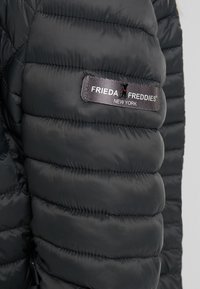Nærbillede af en mørk, fyldig jakkeærme, der viser "Frieda Freddies New York" logoet på et rektangulært skilt. Texturdetaljer i stoffet er synlige.