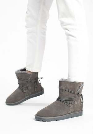 Bottes chelsea en daim gris avec une doublure en fourrure douce, dotées d'une semelle flexible et d'un détail de laçage décoratif. Portées avec un jean slim blanc.