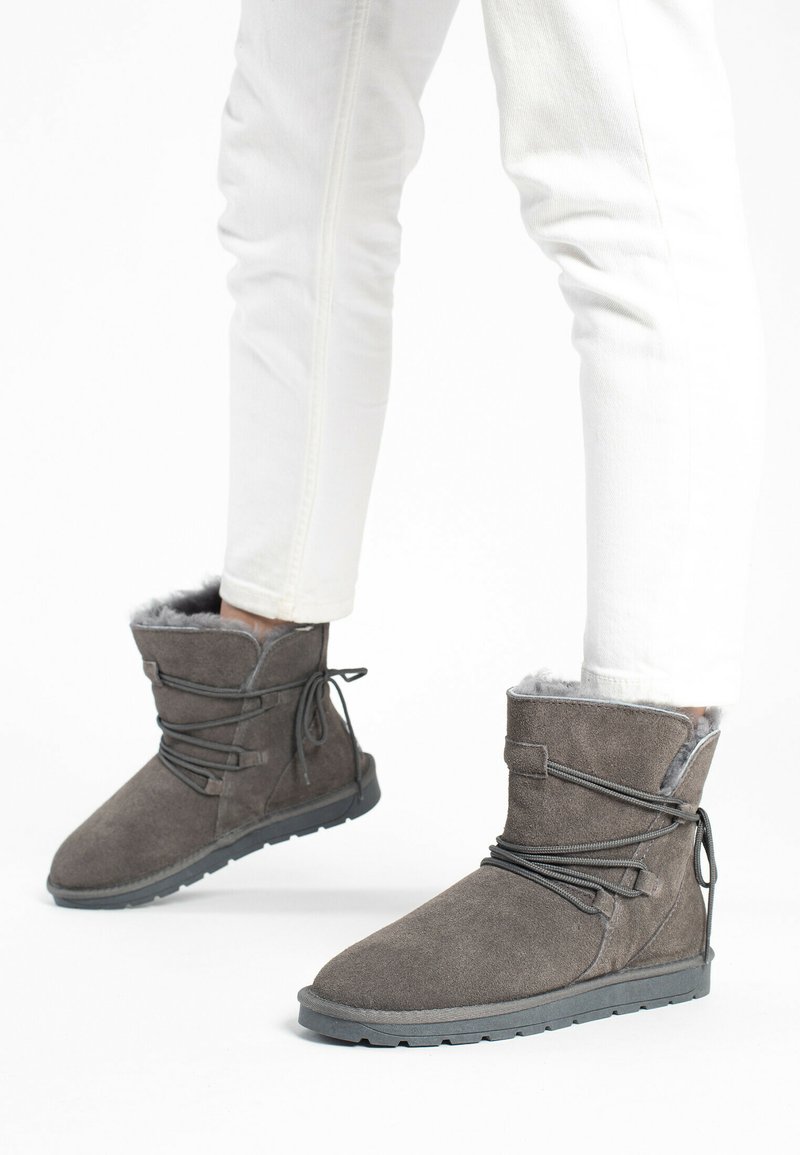 Grå ruskinds ankelstøvler med blød pelsfor, der har en fleksibel sål og dekorativ snøredetalje. Båret med hvide slim-fit jeans.