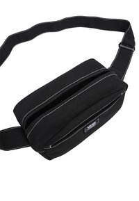 Marsupio nero in tela con due scomparti con zip, dettagli in cucitura bianca e strap regolabile. Presenta una toppa con logo sul davanti.
