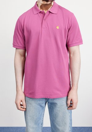 Mężczyzna w jasnoróżowej koszulce polo z krótkim rękawem z małym żółtym logo na lewej piersi, w połączeniu z jasnoniebieskimi dżinsami.