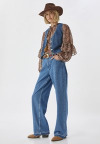 Jeans a gamba larga in denim chiaro abbinati a una blusa paisley marrone e un gilet in denim blu, completati da un cappello marrone fondente e stivaletti alla caviglia.