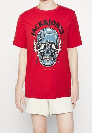 T-shirt rouge avec un grand graphisme de crâne robotique bleu, texte "JACK & JONES" au-dessus, porté avec un short beige clair sur une personne, vue de face.