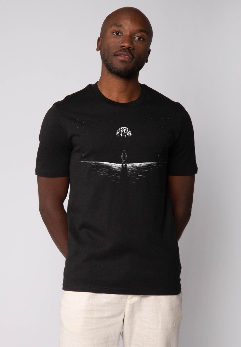 T-shirt en coton noir à manches courtes et encolure ronde. Présente un design graphique blanc d'une silhouette se tenant près de l'eau, sous une forme en dôme.