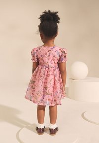 Roosa lilleline kleit puffy varrukatega, kogutud vöökohaga ja nööpide detailiga. Seljas valged volangidega sokid ja läikivad pruunid kingad. Tagantvaade.