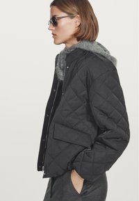Black quilted bomber jacket with a gray fur-lined hood, featuring a zip closure and side pocket, worn with gray pants and sunglasses.