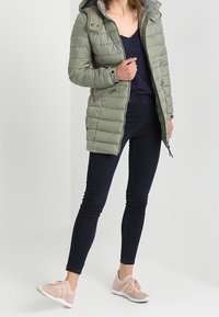 Chaqueta larga acolchada verde oliva con capucha, cierre de cremallera y dos bolsillos, combinada con jeans oscuros ajustados y zapatillas ligeras de color rosa.