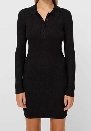 Femme portant une robe en maille côtelée noire, ajustée, à manches longues, avec un col et une patte à quatre boutons, debout devant un fond uni.