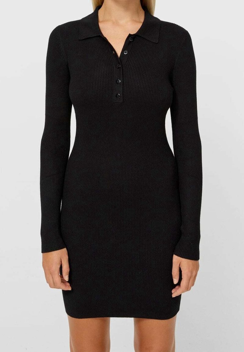 Femme portant une robe en maille côtelée noire, ajustée, à manches longues, avec un col et une patte à quatre boutons, debout devant un fond uni.