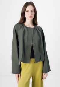 Veste cropped vert olive avec un encolure froncée, fermeture à bouton et deux poches avant ; associée à un pantalon large texturé jaune.
