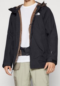 Chaqueta negra impermeable con interior marrón, con cierre frontal, capucha ajustable, puños elásticos y bolsillos laterales. Llevada sobre una camiseta blanca.