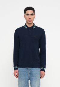 Navy long-sleeve polo shirt with green and white striped collar, button placket, and embroidered logo detail on the chest. Worn with blue jeans.