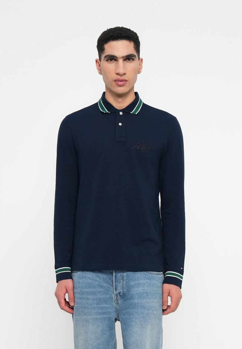 Navy long-sleeve polo shirt with green and white striped collar, button placket, and embroidered logo detail on the chest. Worn with blue jeans.