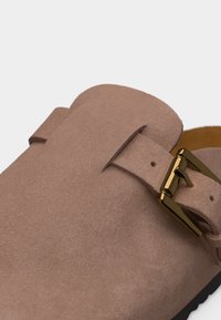 Καφέ καστόρινο παπούτσι τύπου slip-on με χρυσή μεταλλική αγκράφα στο πάνω μπροστινό μέρος.