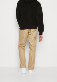 Beige Hose mit schmaler Passform, ausgestattet mit einer Gesäßtasche und einem subtilen Markenetikett, kombiniert mit einem schwarzen, strukturierten Fleece-Hoodie.