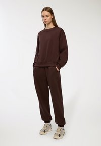 Brązowy bawełniany sweter i pasujące spodnie z elastycznymi mankietami. Model w trampkach, stojący na jasnoszarym tle.