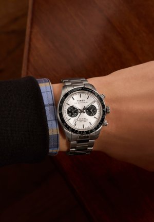Edelstahl-Chronographenuhr mit weißem Zifferblatt, schwarzem Lünette und zwei schwarzen Unterziffernblättern. Getragen an einem Handgelenk mit einem schwarz-blauen karierten Ärmel.
