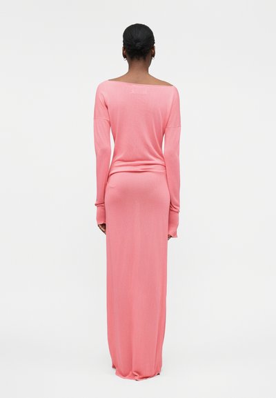 Γυναίκα με σκούρα μαλλιά σε χαμηλή σινιόν, που φορά ένα μακρύ, εφαρμοστό, ροζ φόρεμα χωρίς ώμους, στέκεται με την πλάτη στην κάμερα σε λευκό φόντο.