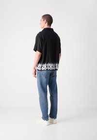 Cămașă neagră cu mânecă scurtă, cu text grafic alb la tiv, purtată cu blugi din denim albastru și adidași albi. Material neted, croi lejer.