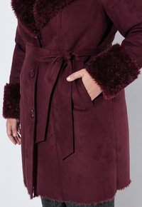 Burgundy kappa i mjukt material med en stor krage och manschetter av päls, med ett bälte och knäppning fram. Strukturerad fåll och sidoficka.