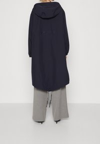 Manteau long imperméable bleu marine avec capuche, poignets élastiques, ourlet à cordon de serrage et un branding subtil dans le dos ; associé à un pantalon large gris clair.