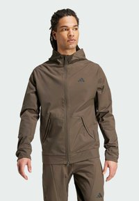 Brun zip-up jakke med hætte, lavet af letvægtsmateriale; har lynlåslommer og et diskret sort logo på brystet.