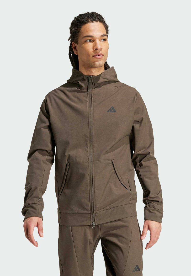 Brun zip-up jakke med hætte, lavet af letvægtsmateriale; har lynlåslommer og et diskret sort logo på brystet.