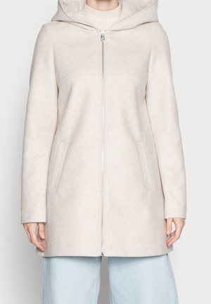 Frau trägt einen hellbeigen Kapuzenmantel mit Frontreißverschluss und seitlichen Taschen über hellblauen Jeans.